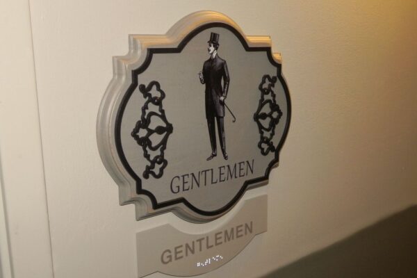 A sign on a wall indicating a gentlemen's restroom. The sign features a vintage-style illustration of a man in formal attire with a top hat and cane, and the word "GENTLEMEN" written below the illustration. The word is also repeated in braille at the bottom.