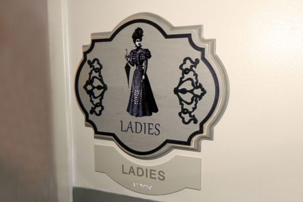 A restroom sign for ladies featuring an illustration of a Victorian-style woman in a long dress with a parasol. The sign has a decorative border and the word "LADIES" appears below the illustration.