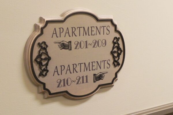 A decorative sign hanging on a beige wall points to apartment numbers 201-209 on the left and 210-211 on the right. The sign is ornate with intricate border detailing and black text.