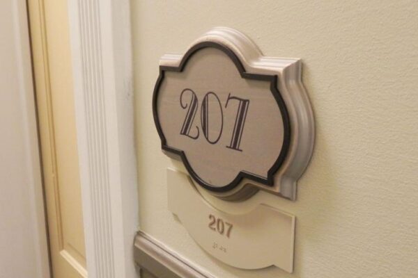 A decorative sign with the number "207" is mounted on a beige wall next to a door. Below the sign is a smaller plaque also displaying the number "207." The background includes door trim details in a light color.