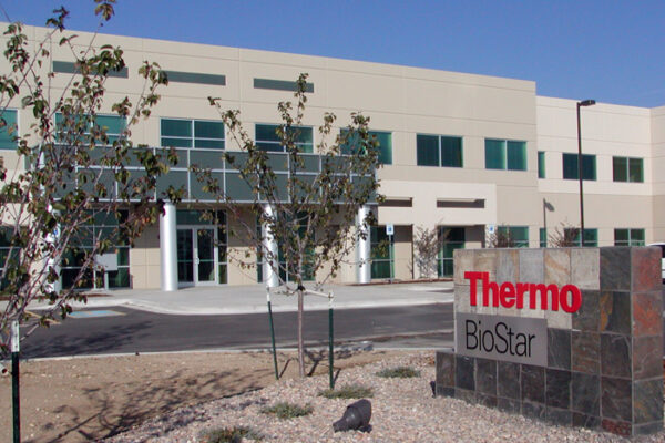 A modern, two-story commercial building with large windows and a sign in front reading "Thermo BioStar" in red and black letters. The surrounding area has young trees and a gravel landscape.
