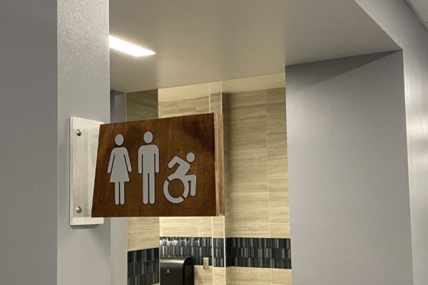 A restroom sign mounted on a light-colored tiled wall depicts icons for male and female restrooms and an additional icon indicating wheelchair accessibility.
