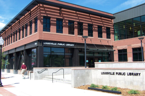 A two-story brick building with large windows showcases the Louisville Public Library. The modern structure features a sign displaying its name on a low concrete wall in front. A person is walking near the entrance under a sunny blue sky.