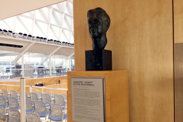 A bronze bust is displayed on a wooden pedestal next to an informational plaque. In the background, there is a bright, spacious atrium with rows of empty chairs arranged in a semicircle, large windows, and a modern architectural design.