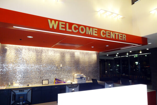 A modern welcome center with a bright red sign reading "Welcome Center" in bold white letters. The desk area below is well-lit and equipped with a computer, a printer, and other office supplies. The background features a textured metallic wall.