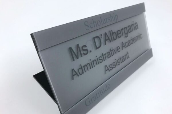 A gray desk nameplate with black lettering reads "Ms. D'Albergaria, Administrative Academic Assistant." The word "Scholarship" is engraved at the top, and "Gratitude" is engraved at the bottom.