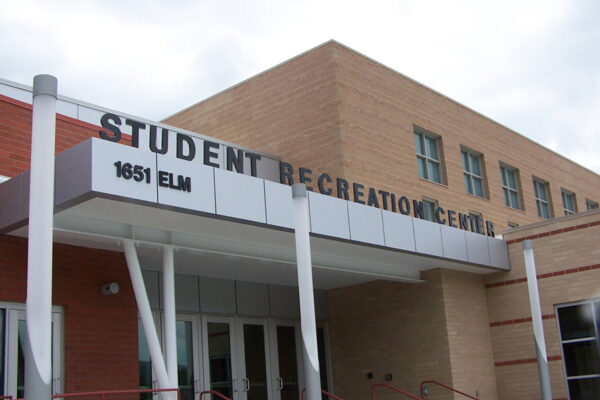 A brick building with a metal awning over an entrance reads "Student Recreation Center" with the address "1651 Elm" displayed prominently. The entrance features glass doors, and there is a red handrail along a short stairway leading to the door.