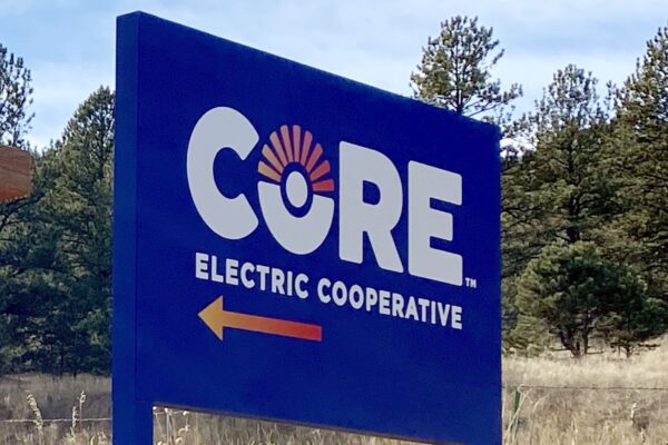 A large blue sign with the words "CORE Electric Cooperative" in white lettering and a circular logo resembling a sun. An orange arrow points to the left beneath the text. The sign is situated in a grassy area with trees in the background.