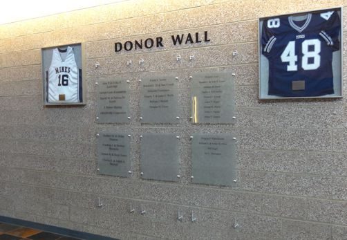 A "Donor Wall" display on a textured wall features framed jerseys on either side, marked with the numbers 16 and 48. Below the sign are several plaques listing donor names and contributions. The plaques are arranged in a grid with clear glass covers.