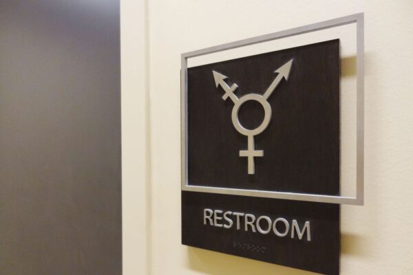 A sign on a wall denotes a restroom with a symbol combining male, female, and non-binary gender symbols. The word "Restroom" is written below the symbol.
