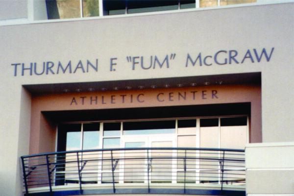 The image shows the exterior of the Thurman E "Fum" McGraw Athletic Center. The name is prominently displayed above a balcony with metal railings. The structure is modern with a neutral color palette.
