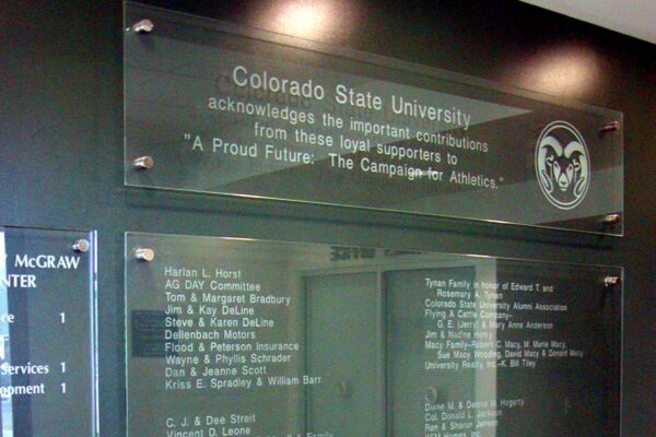 Glass donor recognition display at Colorado State University, showcasing names of individual and corporate benefactors. Etched text includes lists of contributors divided into sections, with the university logo visible at the top right corner.