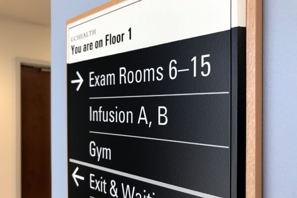 A directional sign inside a healthcare facility indicates that on Floor 1, Exam Rooms 6-15, Infusion A and B, and the Gym are to the right. The Exit & Waiting area, Toilets, and Exam Rooms 1-5 are to the left.