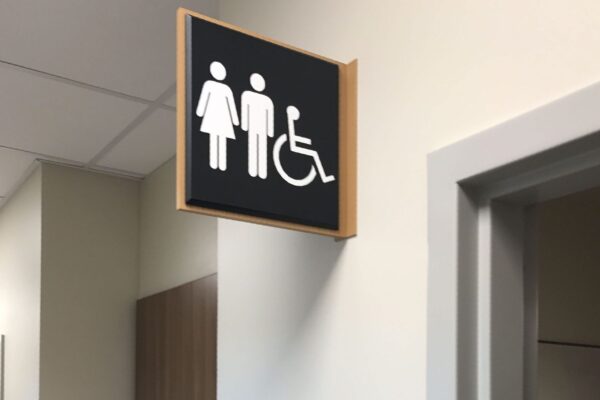 A restroom sign is mounted above a hallway, indicating a unisex toilet with accessibility features. Below it is another sign on the wall with symbols for male, female, and accessible, accompanied by the word "TOILET." A sink is visible in the background.