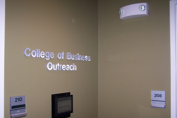 Signs on a beige wall indicate "College of Business Outreach." Additional signs show a room labeled 210, featuring a display screen beneath the sign, and another room labeled 208. A ceiling light fixture is visible above.