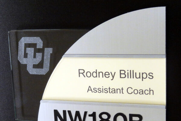 Office nameplate on a black background. The nameplate has a frosted panel with the letters "CU", a metal plate below it with "Rodney Billups," "Assistant Coach," and a bottom metal panel with "NW180B" in raised, black letters and Braille beneath the text.