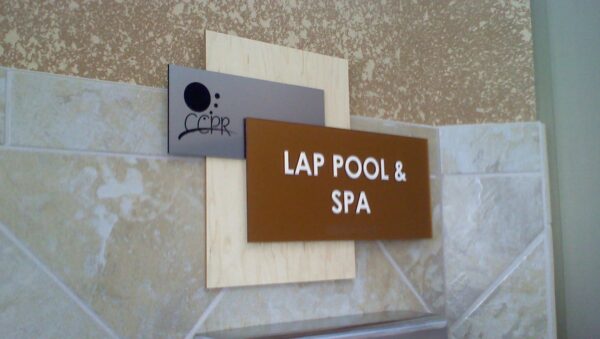 Signage on a tiled wall indicates the direction of the lap pool and spa. The sign has a brown rectangular section with white text that reads "LAP POOL & SPA" and a smaller grey square section on top with an abstract design and the word "CCPR".