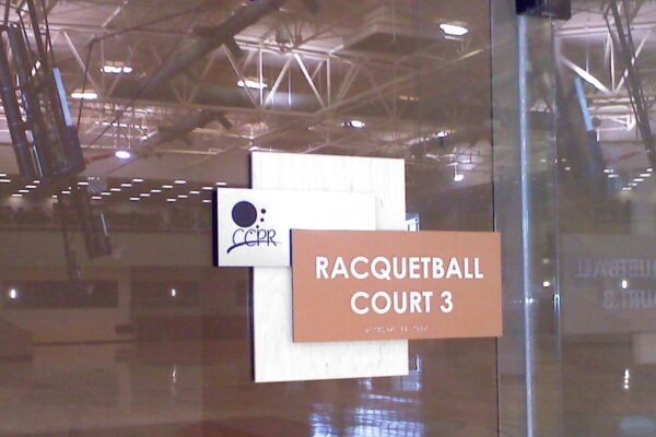 A glass door with a sign reading "Racquetball Court 3" in white text on a brown background. The interior of the racquetball court is visible through the glass door, showing the court surrounded by transparent walls and a ceiling with visible lights and beams.
