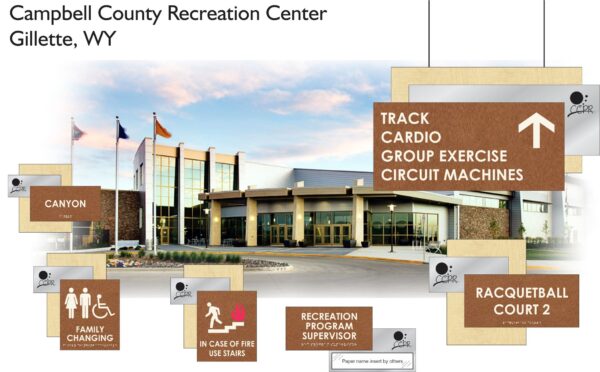 Exterior view of the Campbell County Recreation Center in Gillette, WY, with signs indicating various facilities such as a track, cardio area, exercise circuit machines, racquetball court, canyon, family changing room, and stairs. The background shows a modern building with flags.