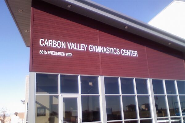 A building with a maroon-colored facade bearing the sign "Carbon Valley Gymnastics Center" in white letters. The address "0615 Frederick Way" is also displayed below the sign. The lower half of the building features large glass windows.