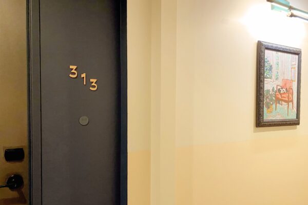 A dark gray door with the number "313" is located on the left side of a beige hallway. To the right of the door, a framed painting depicting a room with a red chair and a table hangs on the wall. The hallway is illuminated by overhead lighting.