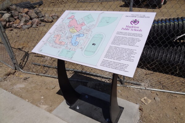 An outdoor informational sign and map on a metal stand for Alpine Public Schools, placed on a concrete slab beside a chain-link fence and construction materials. The sign contains colorful sections and text for visitors to read.