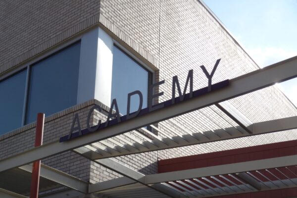 A modern building with large windows has an overhang featuring a sign with the word "ACADEMY" in capital letters. The structure is made of brick and metal, and the sky is clear in the background.