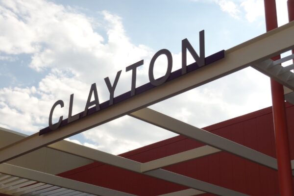 A metal structure with "CLAYTON" displayed in large, capital letters against a partially cloudy sky. The building features a mix of red and beige colors, and the photograph is taken from an upward angle.