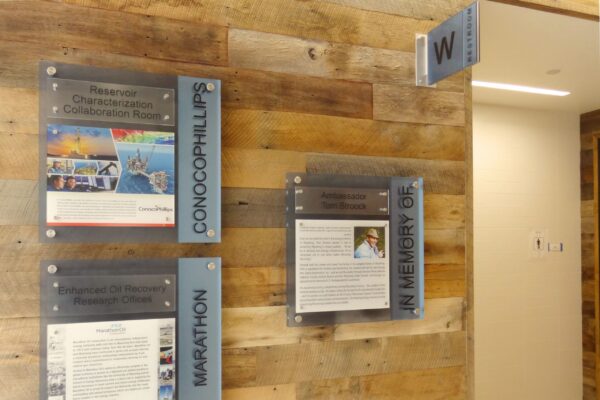 A wooden wall displays three plaques. Two on the left showcase information related to ConocoPhillips and Marathon. The third, titled "In Memory Of," features a photo and text. A sign with the letter "W" hangs at the hallway entrance.