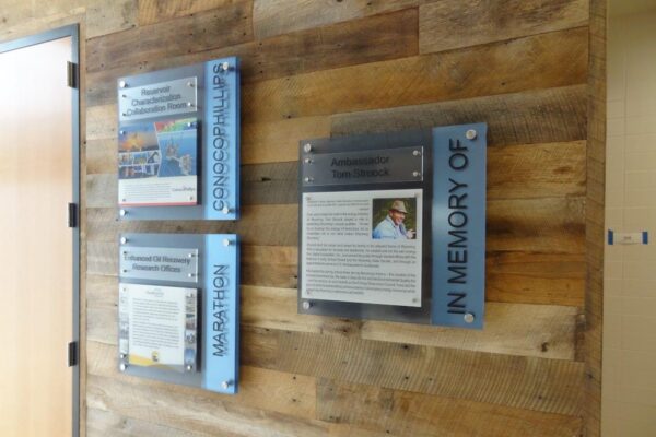 Three plaques are mounted on a wooden wall. They contain text and photos commemorating various subjects. One plaque includes the phrase "IN MEMORY OF." Each plaque is framed with transparent and colored borders, enhancing their prominence on the rustic surface.