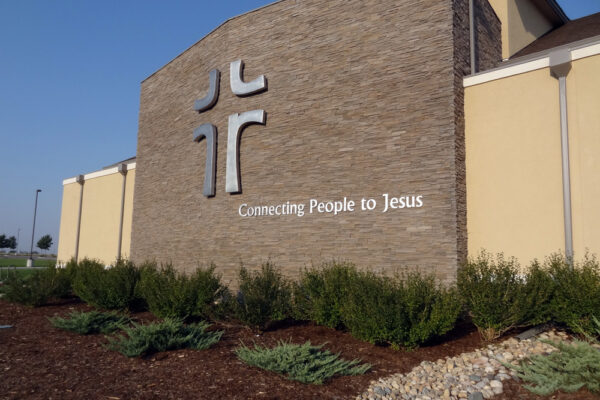 A church building with a stone facade and an emblem resembling crosses on the upper left. Below the emblem, the text "Connecting People to Jesus" is displayed. There are well-maintained bushes and a gravel path in front of the building.