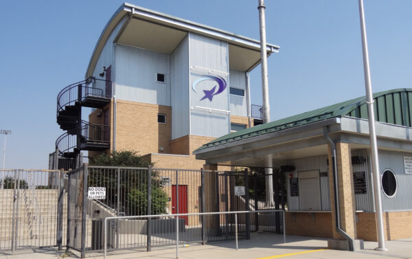A modern building with a logo of a shooting star on it. The structure features a mix of brick and metal siding and includes an outdoor spiral staircase. A security gate and ticketing station are visible in the foreground.