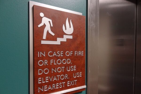 A wall sign next to an elevator depicts a person descending stairs with a flame icon. The text reads: "In case of fire or flood, do not use elevator. Use nearest exit." The sign has a brown background and white text and graphics.