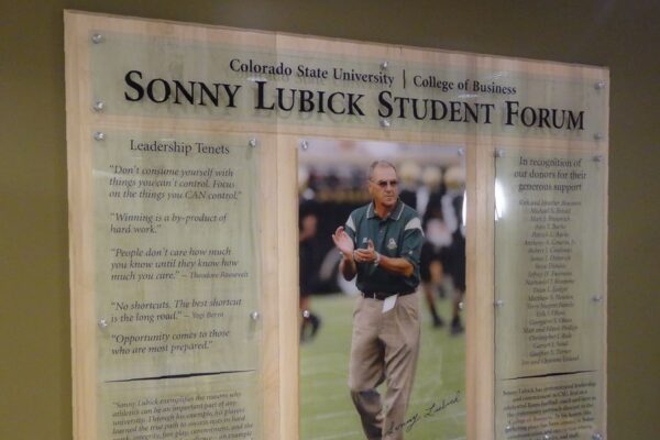 A wall-mounted wooden plaque titled "Sonny Lubick Student Forum" at Colorado State University College of Business, featuring an image of a man in the center with lists of leadership traits on both sides. The plaque highlights leadership qualities and principles.