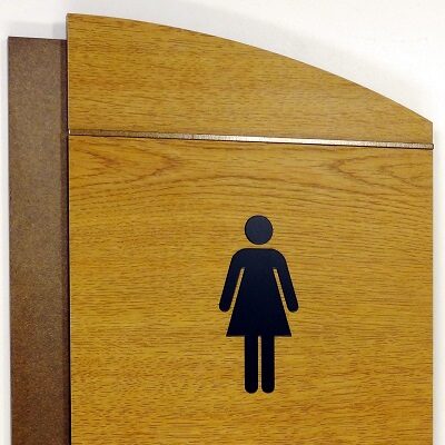 Wooden restroom sign indicating a women's bathroom. The sign has a simple silhouette of a woman and the word "WOMEN" written below in black text. The sign has a rectangular shape with a slightly slanted top.