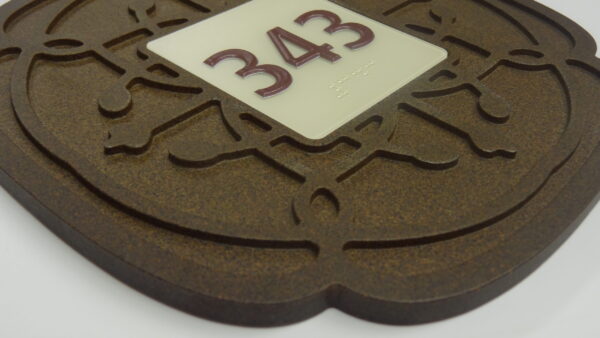 A close-up view of a decorative sign with the number 343 displayed in red on a white background. The sign is mounted on a textured, dark brown base with an intricate design around the edges.