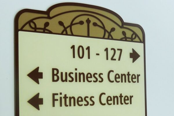 A wall-mounted directional sign with an ornate design points arrows in various directions. To the left are directions to the Business Center, Fitness Center, and Restrooms. An arrow pointing right leads to rooms numbered 101 - 127.