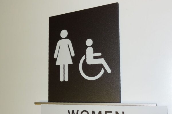 A restroom sign on a wall indicating facilities for women, including those with disabilities. The sign has symbols for a standing woman and a person in a wheelchair above the word "WOMEN". Below the text, there is Braille for accessibility.