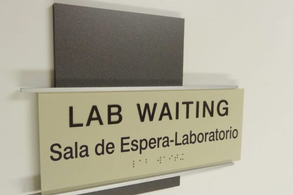 A sign on a wall reading "LAB WAITING" in large text, with "Sala de Espera-Laboratorio" written in smaller text underneath. There is also Braille text below the written words. The sign is mounted on a flat, dark background.