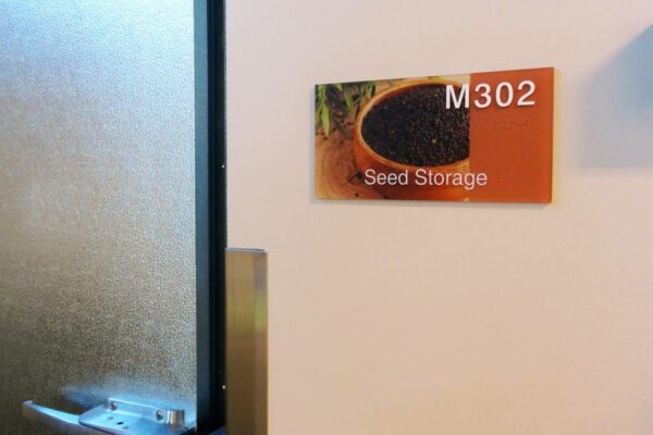 A door with a frosted glass panel is partially open. A sign next to the door reads "M302 Seed Storage" and features an image of a bowl filled with black seeds.
