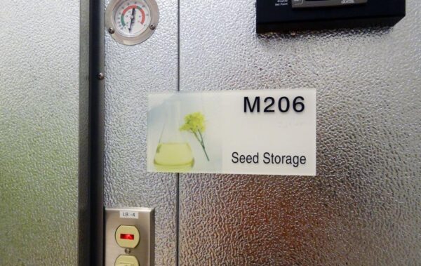 A metal door with a sign reading "M206 Seed Storage." The sign includes an image of a plant and braille writing.