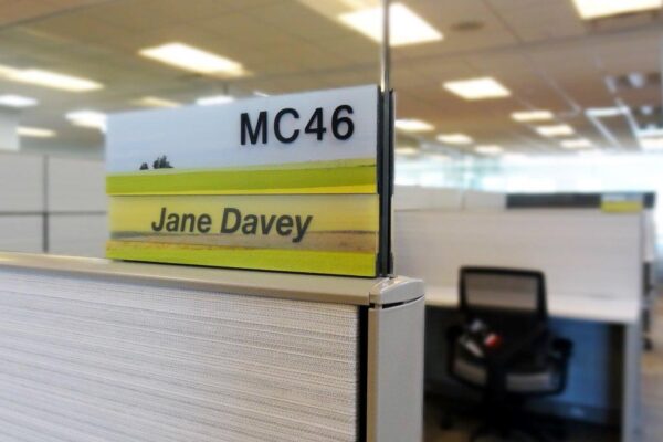 An office cubicle is labeled with a nameplate reading "MC46" and "Jane Davey." The cubicle is empty, featuring a chair and desk inside. The surrounding area includes similar cubicles under fluorescent lighting.