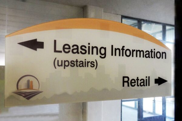 A directional sign indicates Leasing Information is upstairs with an arrow pointing left, and Retail to the right with an arrow pointing right. The sign has a cityscape graphic and is placed indoors near a glass window.