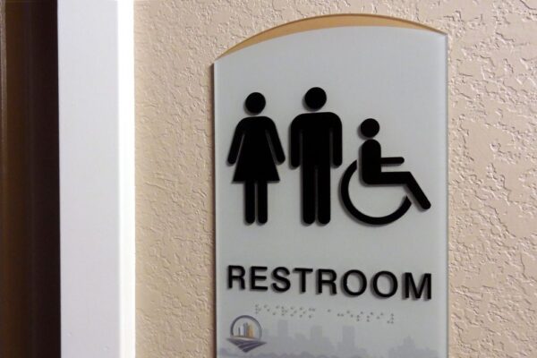 A restroom sign mounted on a wall indicates an accessible bathroom for all genders. The sign shows icons for a woman, a man, and a person in a wheelchair above the word "Restroom." A small logo is visible at the bottom of the sign.