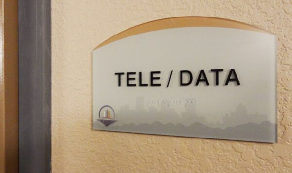 A frosted glass sign on a beige wall reads "TELE / DATA" in bold black letters. Beneath the text, embossed but barely visible, it says “Utilities.” The bottom of the sign features a small, colorful logo.