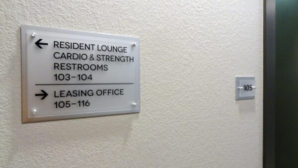 A hallway with a beige wall featuring a sign with black text and arrows. The sign points left for the Resident Lounge, Cardio & Strength, and Restrooms (rooms 103-104), and right for the Leasing Office (rooms 105-116). A door with a room number is partially visible.