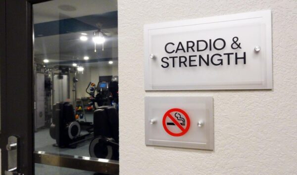 A door with a sign that reads "Cardio & Strength" leads to a gym. Below it, there's a "no smoking" sign. Inside the gym, exercise equipment is visible including a treadmill and elliptical machines. The gym area is lit with ceiling lights.