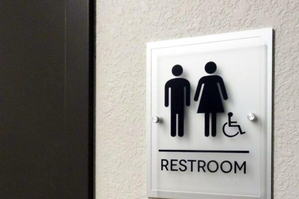 A sign on a wall indicates a restroom. The sign has symbols for a man, a woman, and a person in a wheelchair. The symbols are in black on a white background above the word "RESTROOM." A door with a metallic handle is adjacent to the sign.
