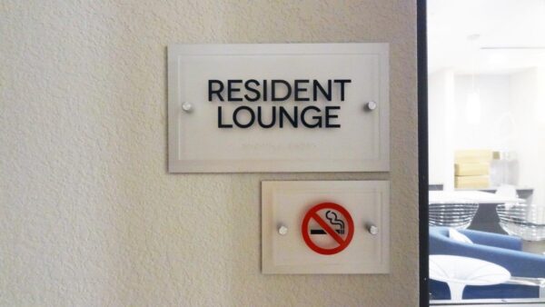 A wall sign reading "Resident Lounge" is displayed with an additional sign below it featuring a no smoking symbol, indicating that smoking is not allowed in the area. The signs are mounted on a light-colored wall near an entrance to the lounge.
