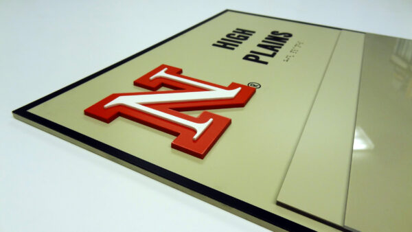 A beige sign with a red "N" logo and the words "High Plains" written in black capital letters. The letters and logo are raised, creating a 3D effect. The right side of the sign appears to have a reflective surface.